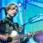 Lindsey Jordan of Snail Mail at the microphone with an acoustic guitar under teal stage light