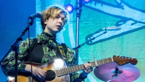 Lindsey Jordan of Snail Mail at the microphone with an acoustic guitar under teal stage light
