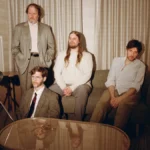 The four members of Protomartyr posed in a hotel room beside a film camera on a tripod