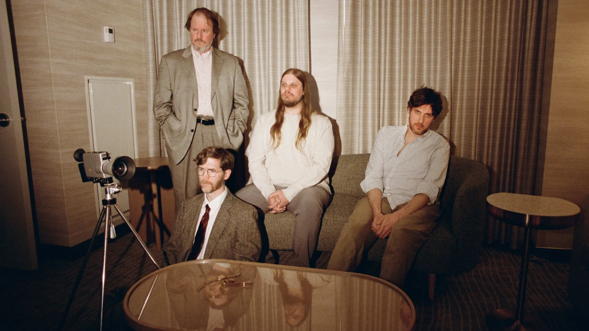 The four members of Protomartyr posed in a hotel room beside a film camera on a tripod